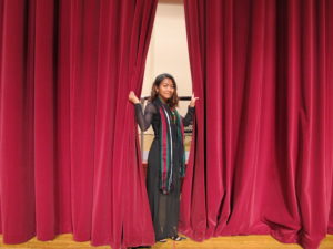 Young lady in a black dress pushing aside a red stage curtain while posing.