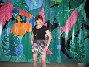 Briél Kate, in a black top and black-and-white striped skirt, posing in front of a colorful wall as part of a selfie photo shoot.