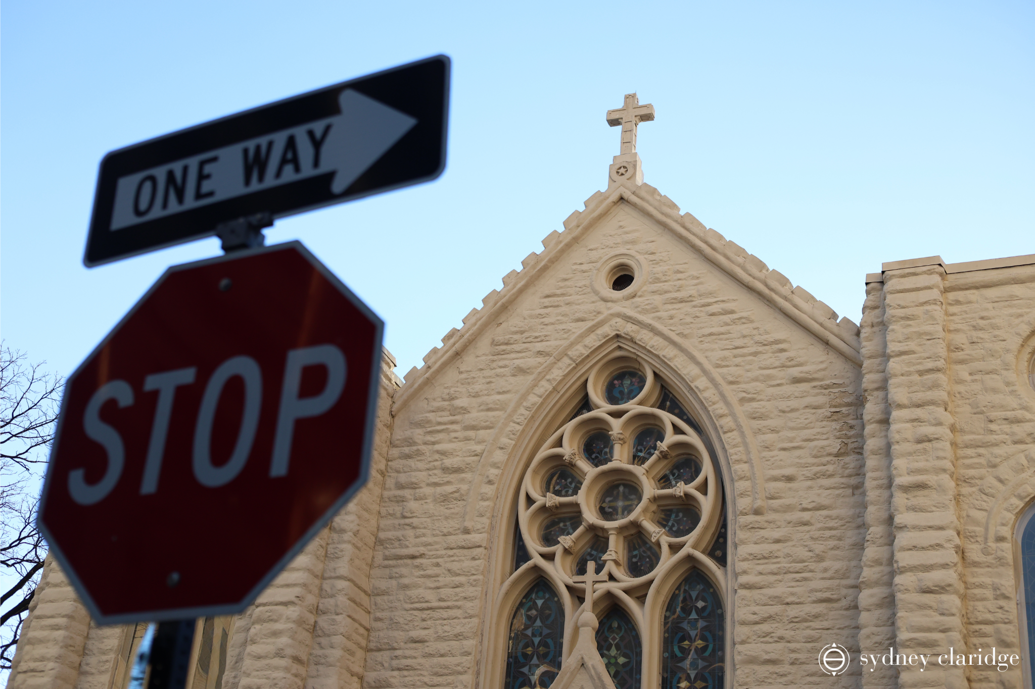 A church is seen behind a stop sign with a one way sign on top. The arrow on the one way sign points to a Christian cross atop the church.