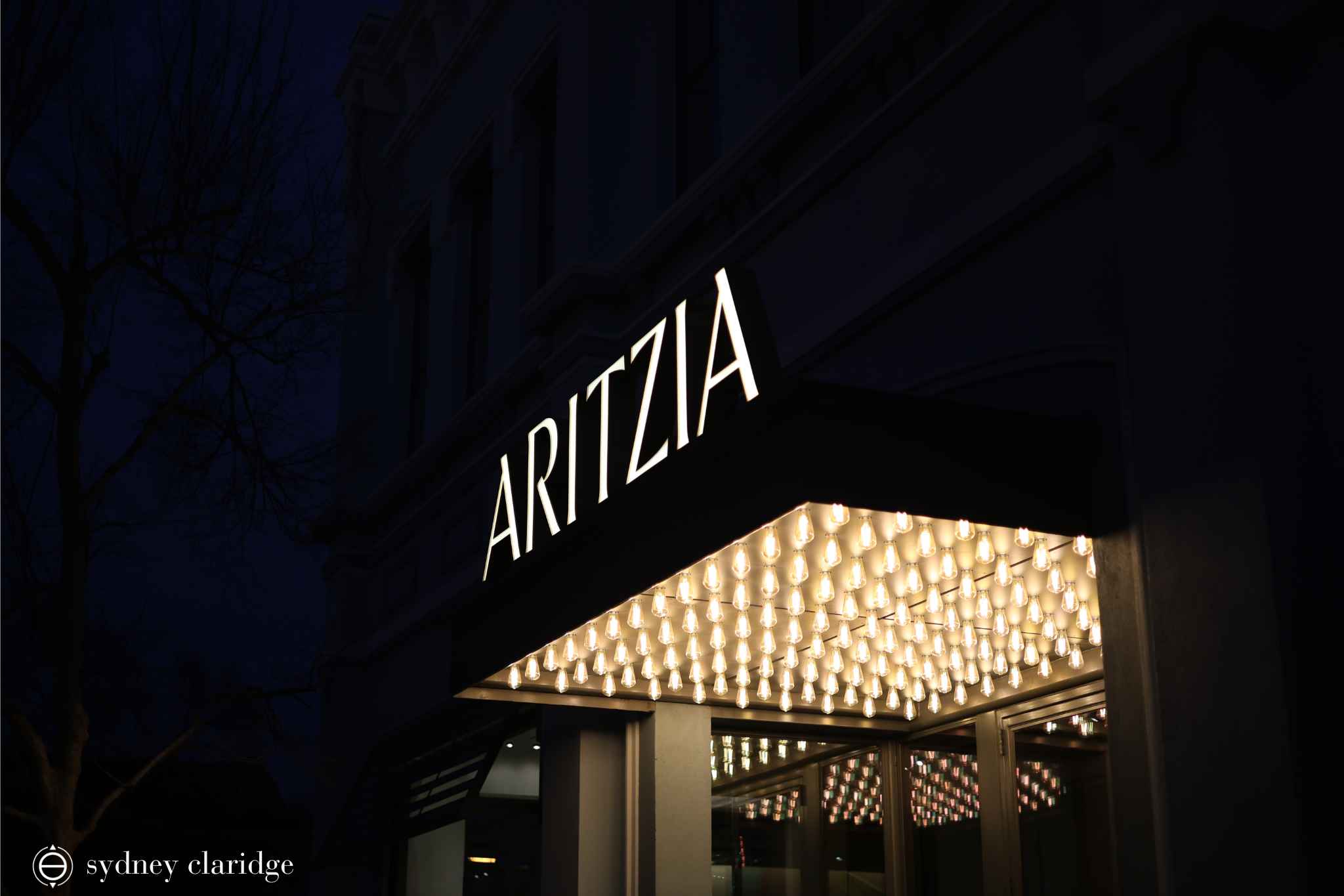 Aritzia store at night. There are multiple lightbulbs under the Aritzia sign.